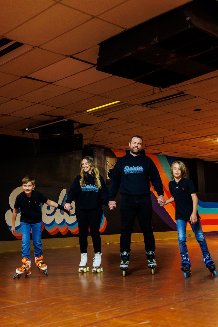A family holding hands and skating together on the Deleta rink floor in front of the rainbow stripe wall